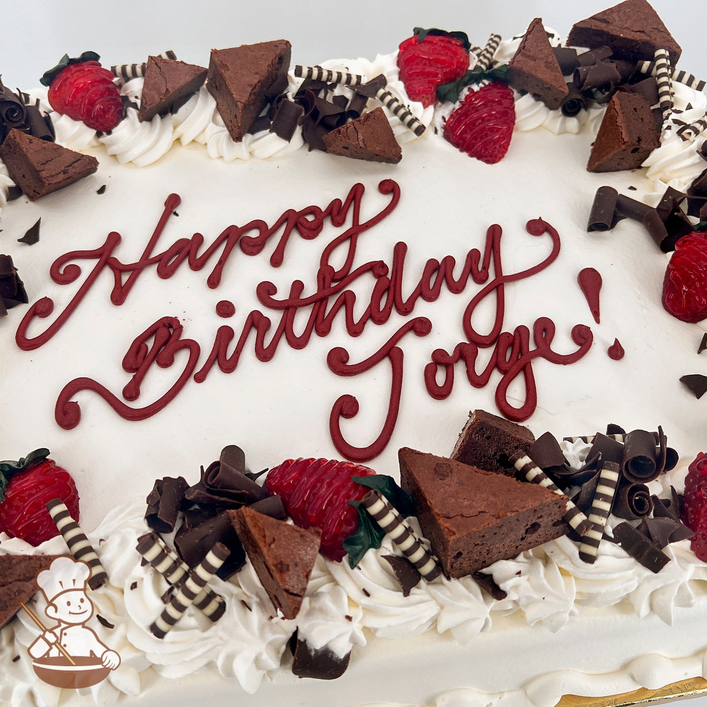 Sheet cake with white frosting, decorated with strawberries, brownie triangles, chocolate curls, and striped chocolate sticks along the edges.