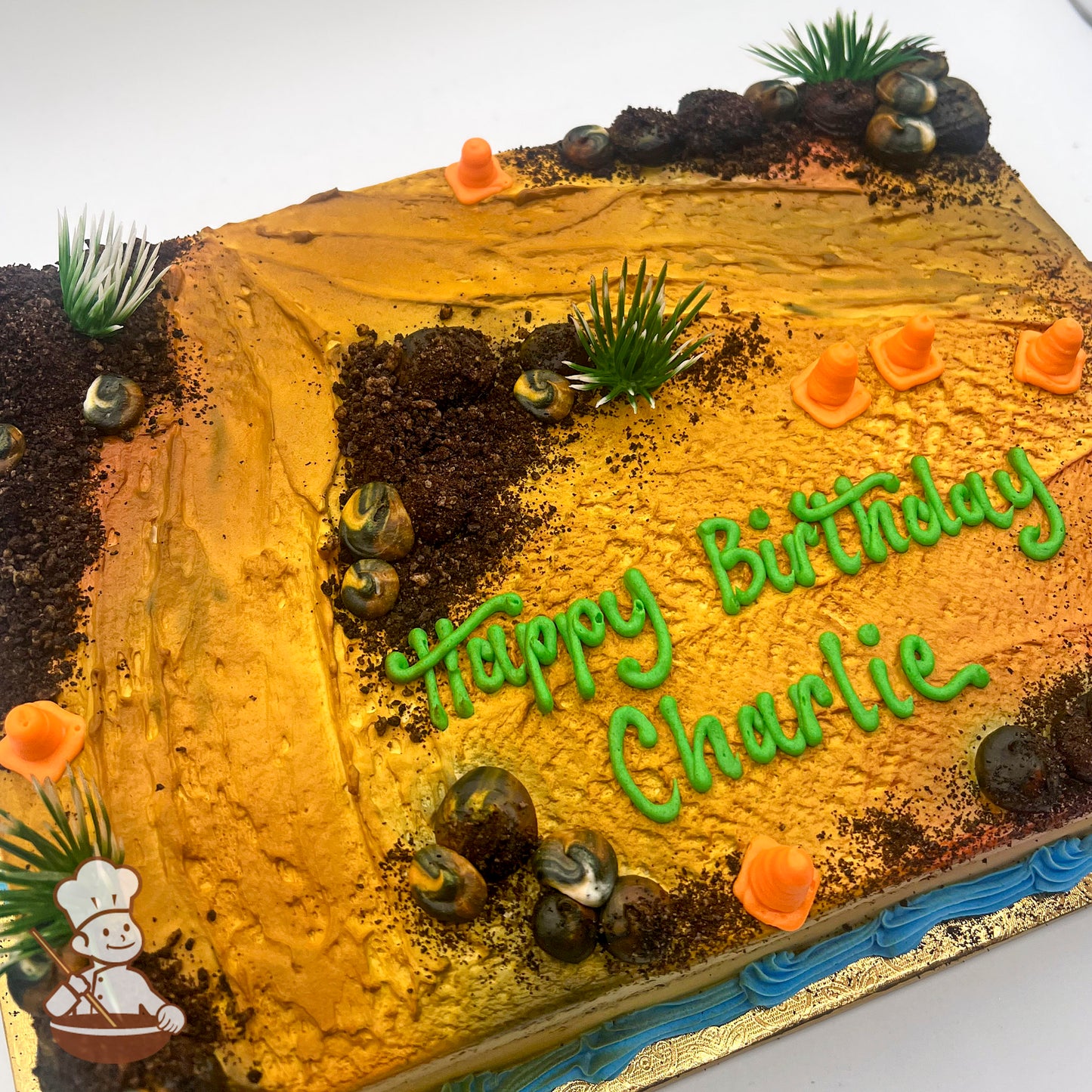 Sheet cake decorated as a construction site with textured golden frosting, cake crumb dirt, plastic bushes, buttercream rocks, and orange cones.