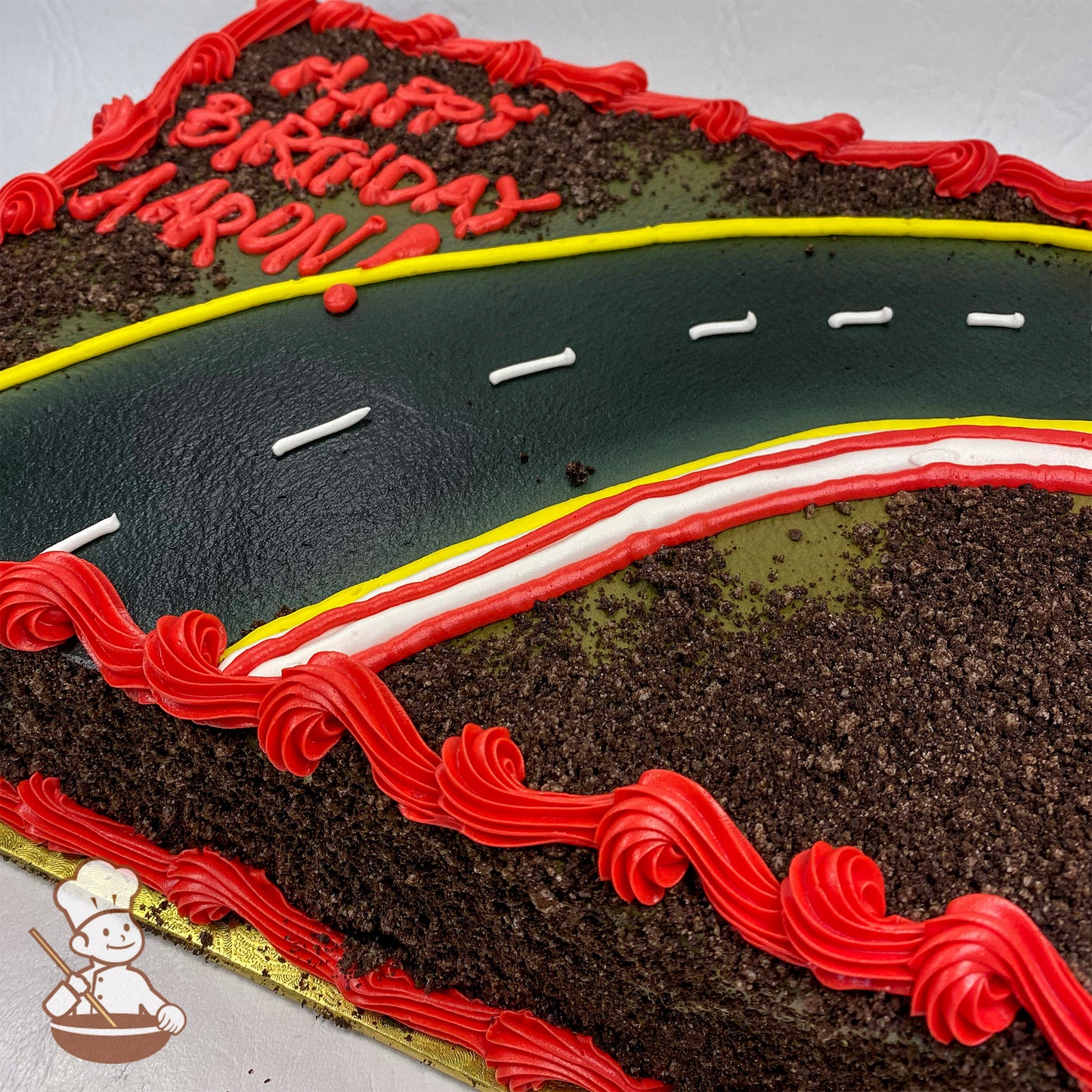 Sheet cake with a curved road design made of black icing, white dashed lines, and yellow and red borders. Crushed cake crumbs resemble dirt, with red buttercream swirl trims along the edges.