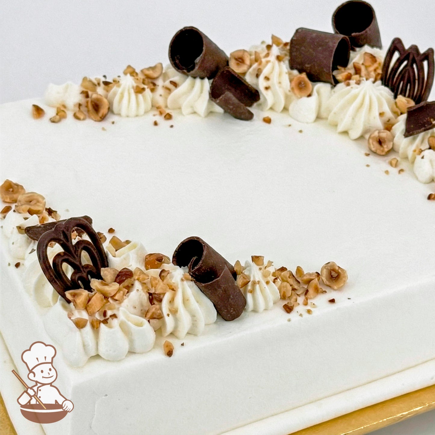 White sheet cake topped with chocolate curls, chocolate shapes, and diced hazelnuts.