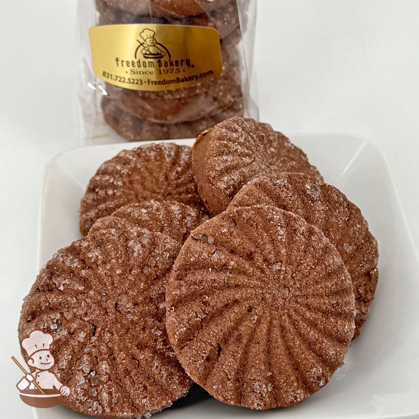 A set of six round, chocolate shortbread cookies with a ridged pattern and sugar crystals on top, displayed on a white plate. A branded bag of cookies is visible in the background.
