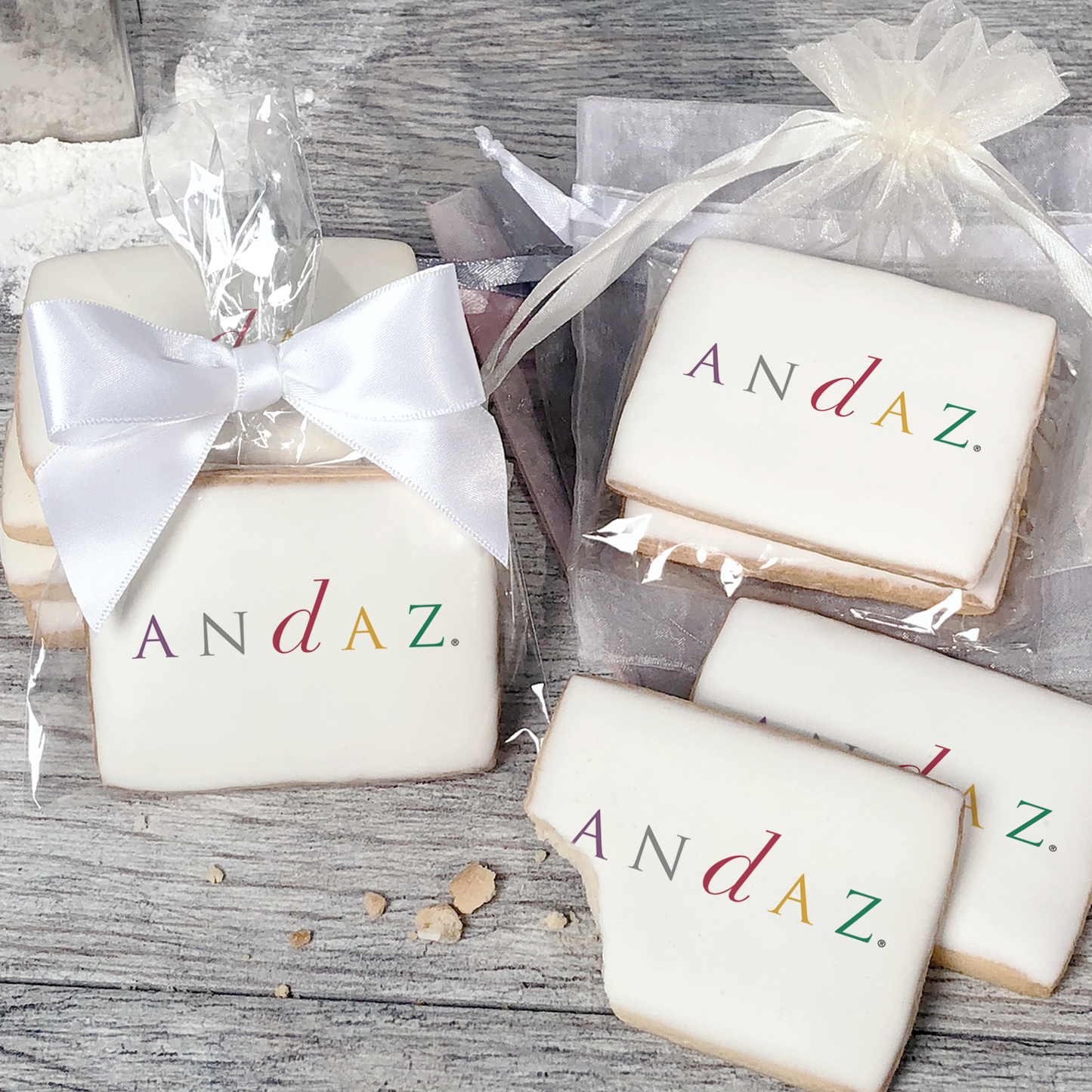 A stack of rectangular butter shortbread cookies with Andaz logo printed directly on a white, light sugar icing. Some cookies are shown in clear packaging with a twist-tie ribbon bow or inside an organza bag.
