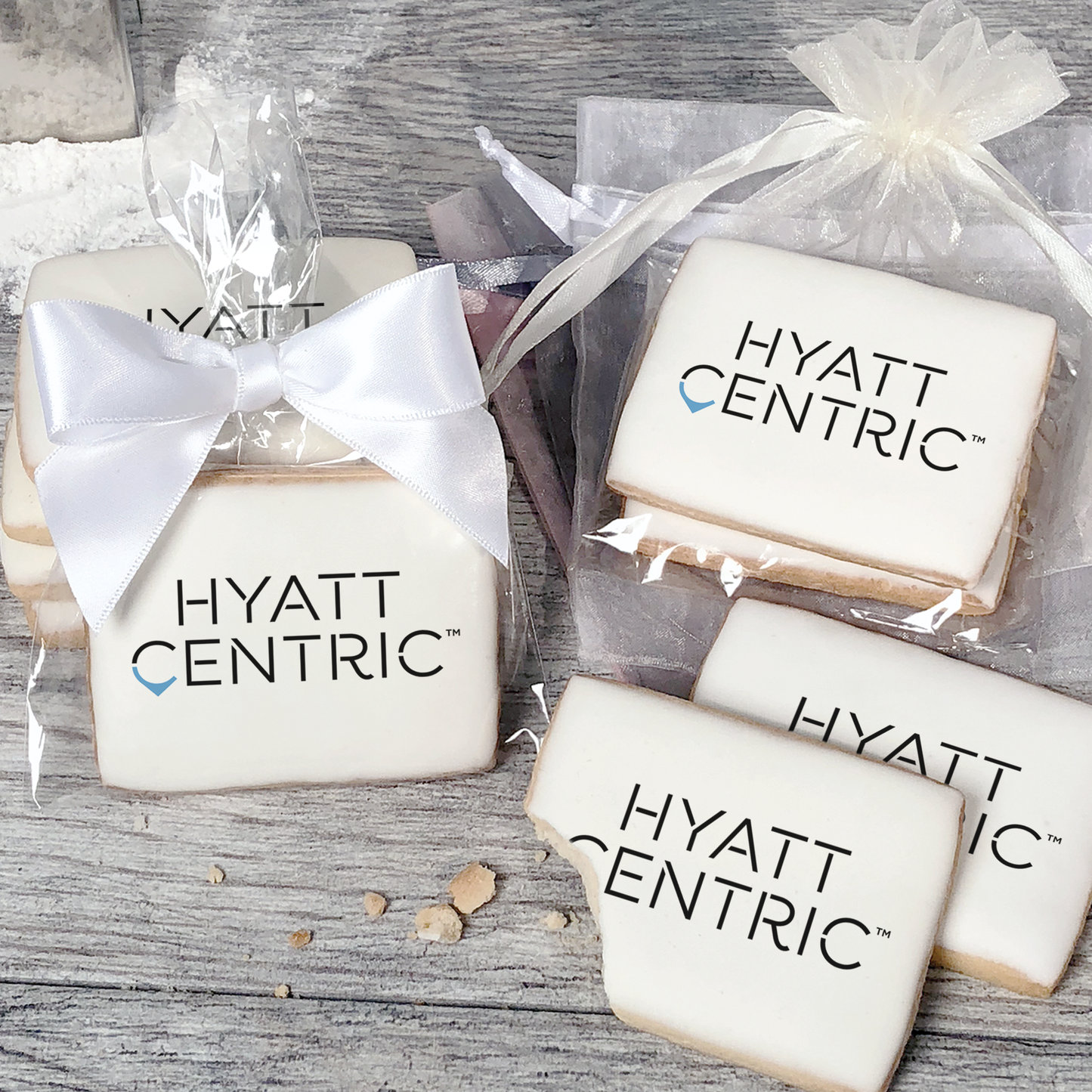 A stack of rectangular butter shortbread cookies with Hyatt Centric logo printed directly on a white, light sugar icing. Some cookies are shown in clear packaging with a twist-tie ribbon bow or inside an organza bag.