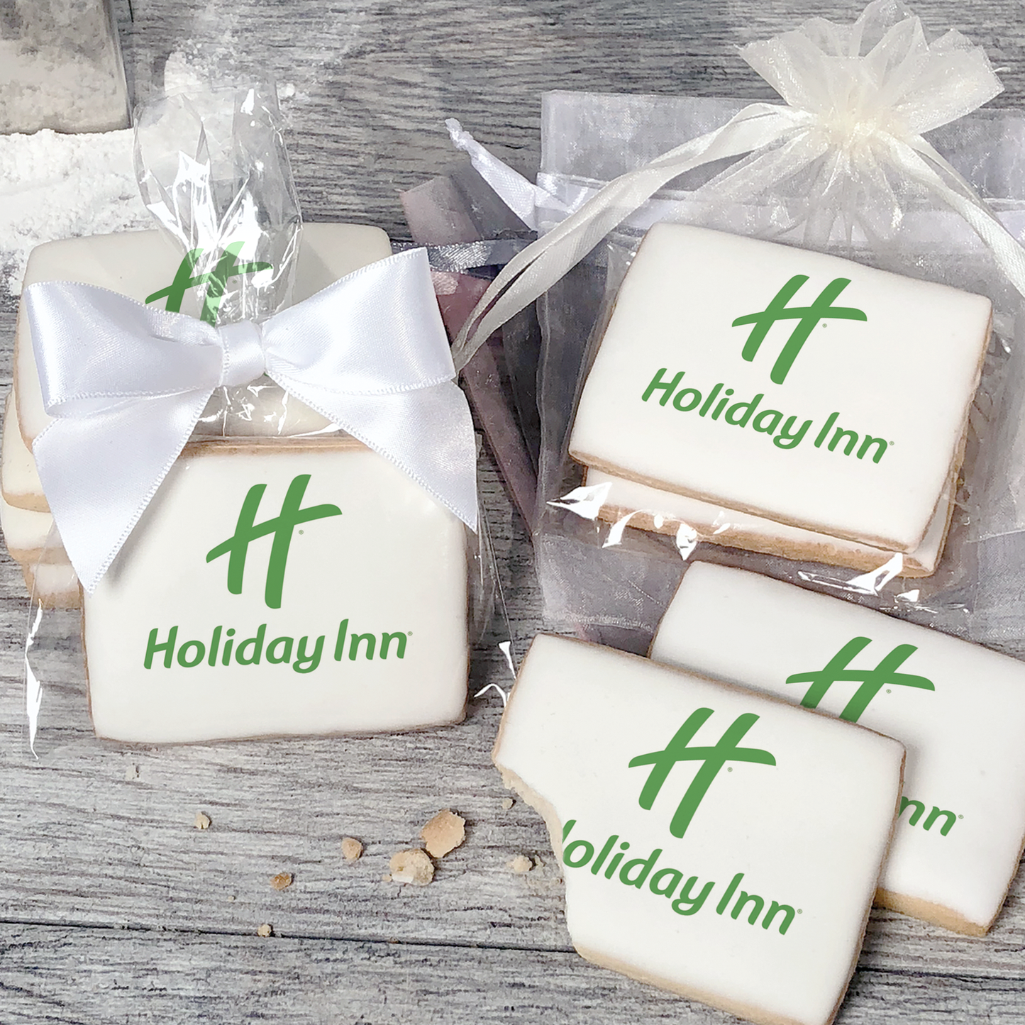 A stack of rectangular butter shortbread cookies with Holiday Inn logo printed directly on a white, light sugar icing. Some cookies are shown in clear packaging with a twist-tie ribbon bow or inside an organza bag.