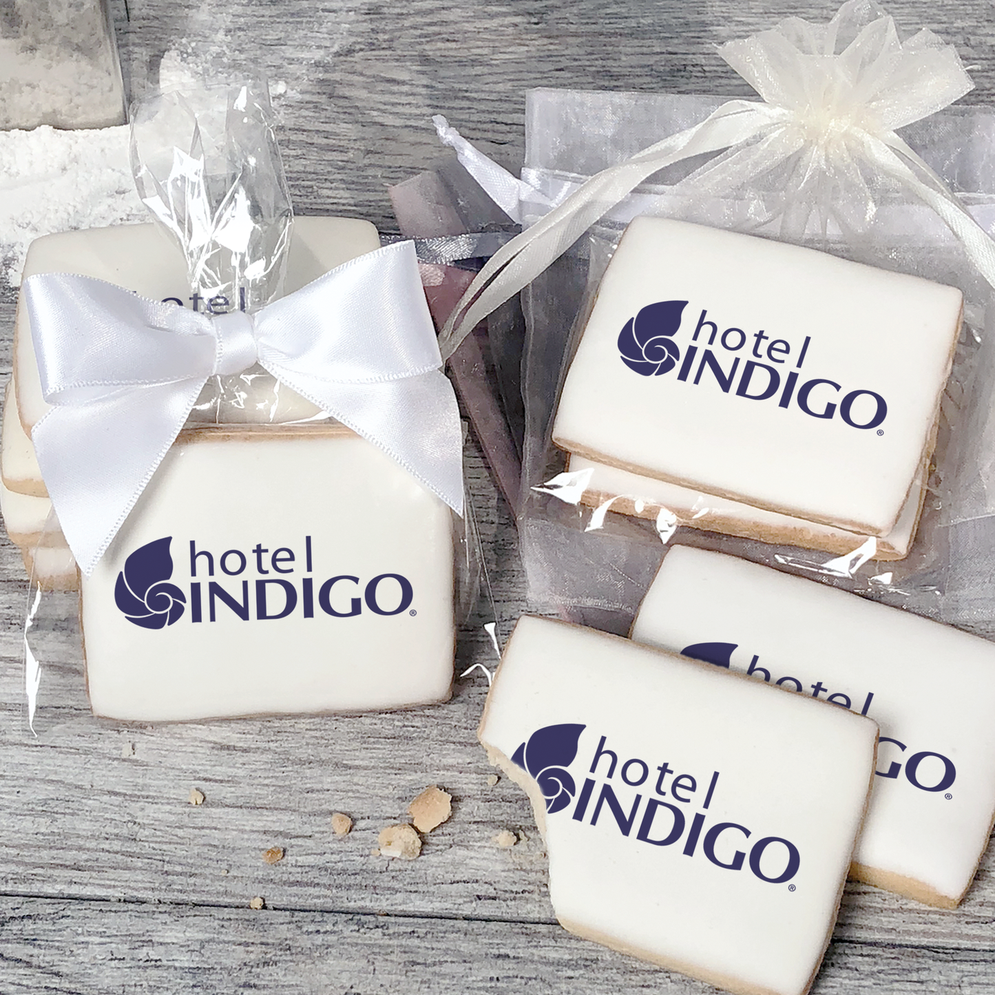 A stack of rectangular butter shortbread cookies with Hotel Indigo logo printed directly on a white, light sugar icing. Some cookies are shown in clear packaging with a twist-tie ribbon bow or inside an organza bag.
