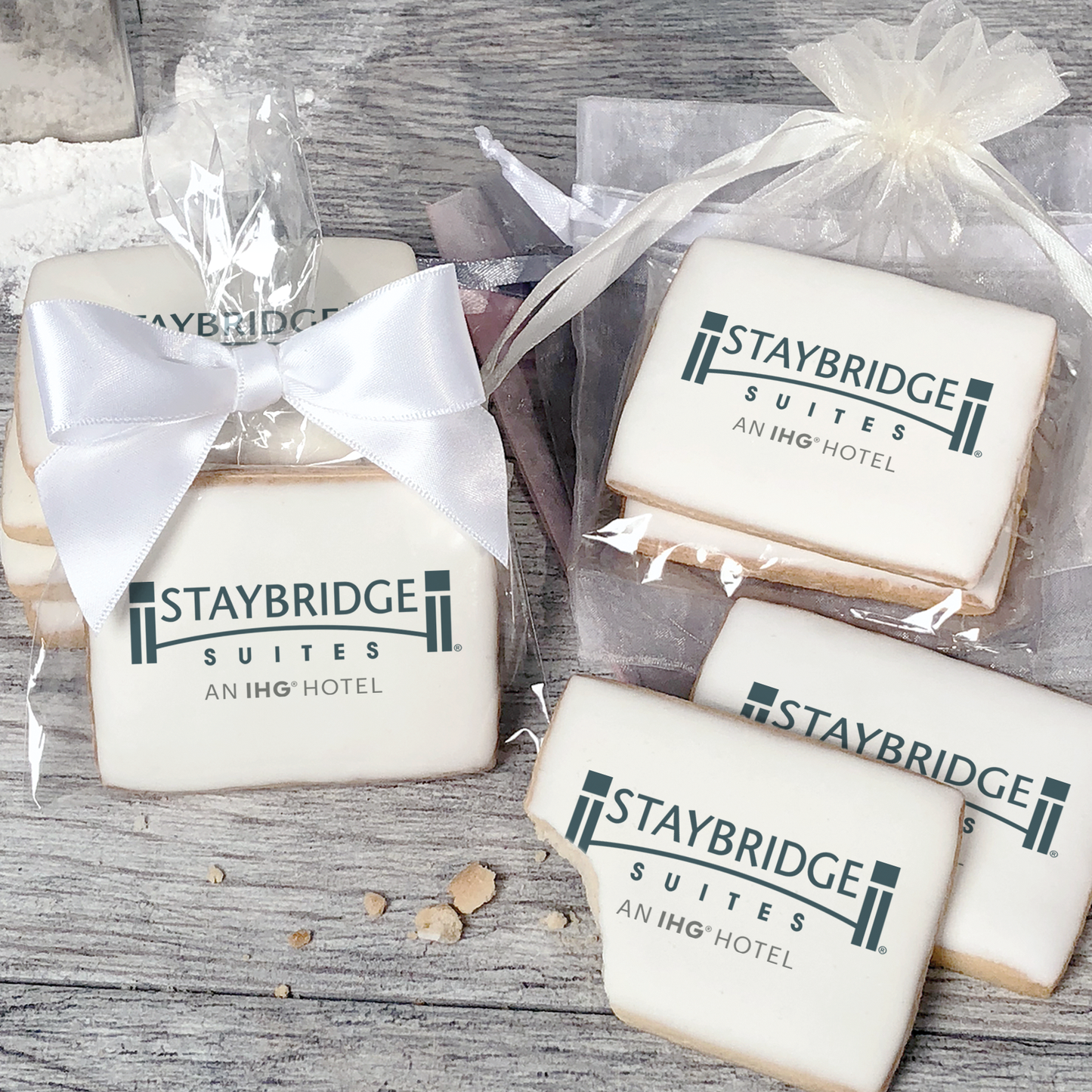 A stack of rectangular butter shortbread cookies with Staybridge Suites logo printed directly on a white, light sugar icing. Some cookies are shown in clear packaging with a twist-tie ribbon bow or inside an organza bag.