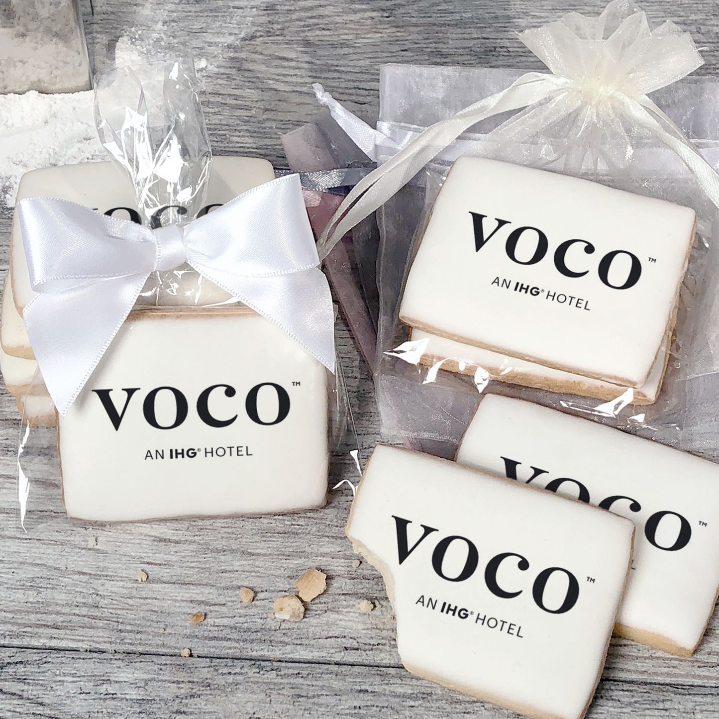 A stack of rectangular butter shortbread cookies with Voco logo printed directly on a white, light sugar icing. Some cookies are shown in clear packaging with a twist-tie ribbon bow or inside an organza bag.