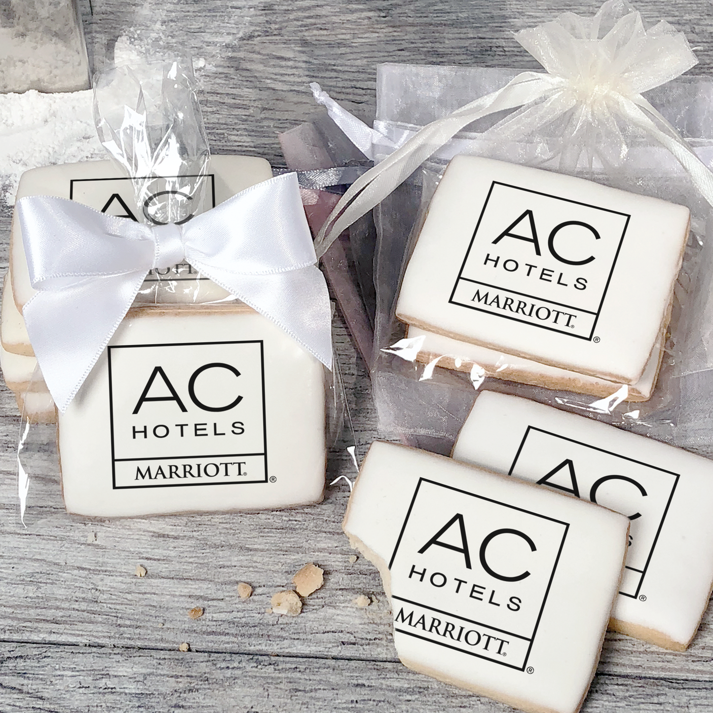 A stack of rectangular butter shortbread cookies with AC Hotels logo printed directly on a white, light sugar icing. Some cookies are shown in clear packaging with a twist-tie ribbon bow or inside an organza bag.