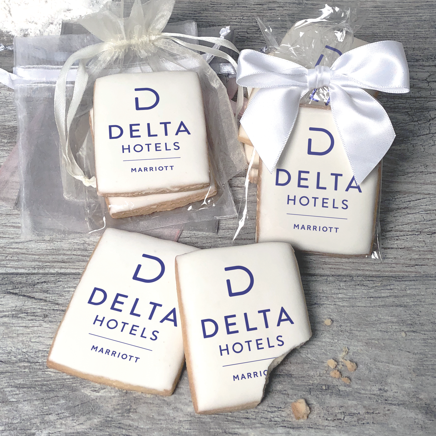 A stack of rectangular butter shortbread cookies with Delta Hotels logo printed directly on a white, light sugar icing. Some cookies are shown in clear packaging with a twist-tie ribbon bow or inside an organza bag.