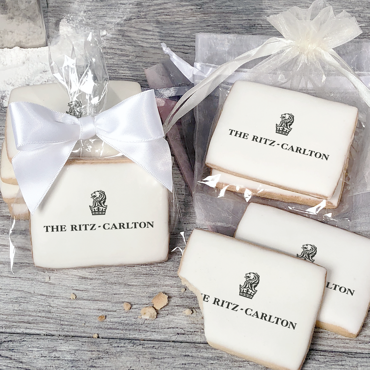 A stack of rectangular butter shortbread cookies with Ritz Carlton logo printed directly on a white, light sugar icing. Some cookies are shown in clear packaging with a twist-tie ribbon bow or inside an organza bag.