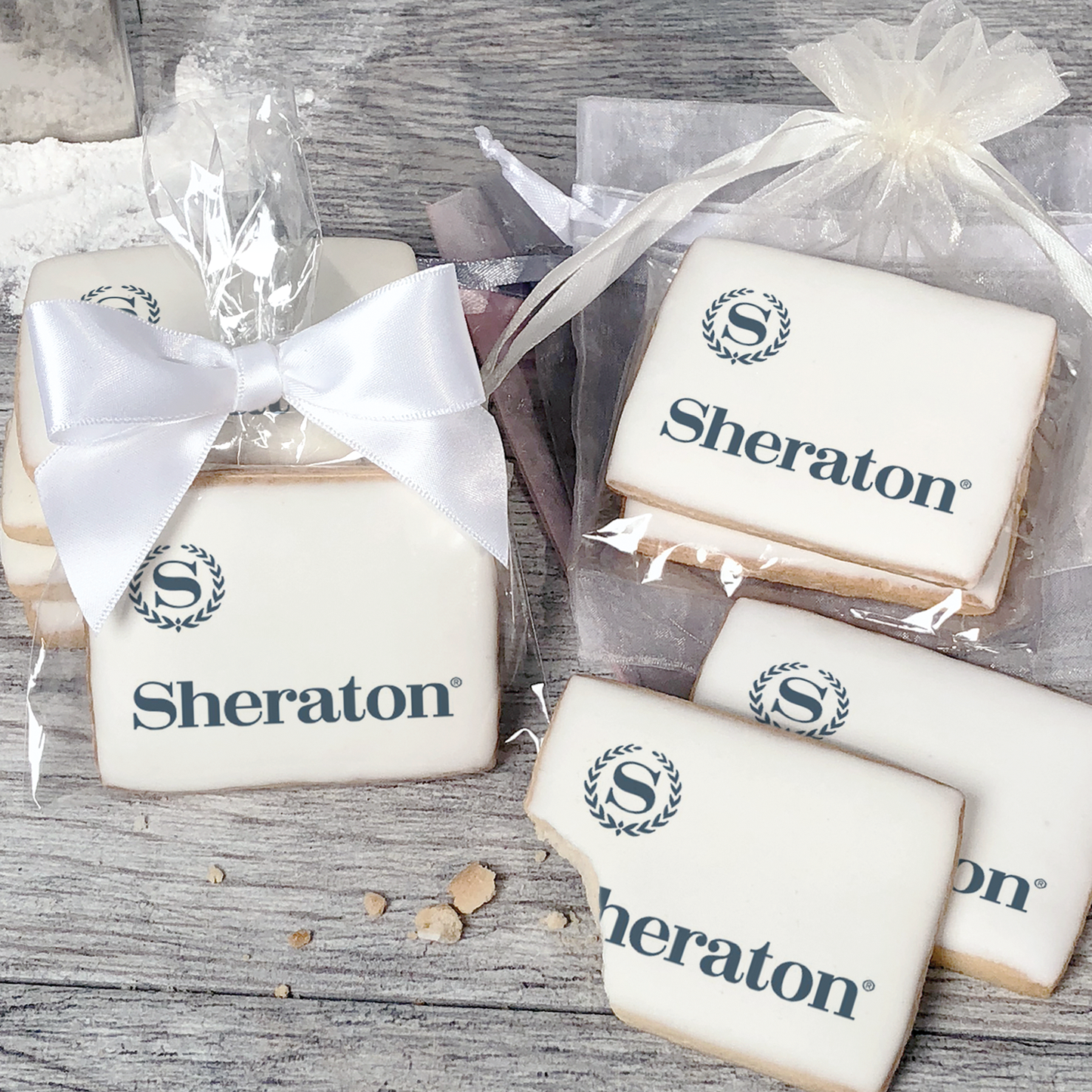A stack of rectangular butter shortbread cookies with Sheraton logo printed directly on a white, light sugar icing. Some cookies are shown in clear packaging with a twist-tie ribbon bow or inside an organza bag.