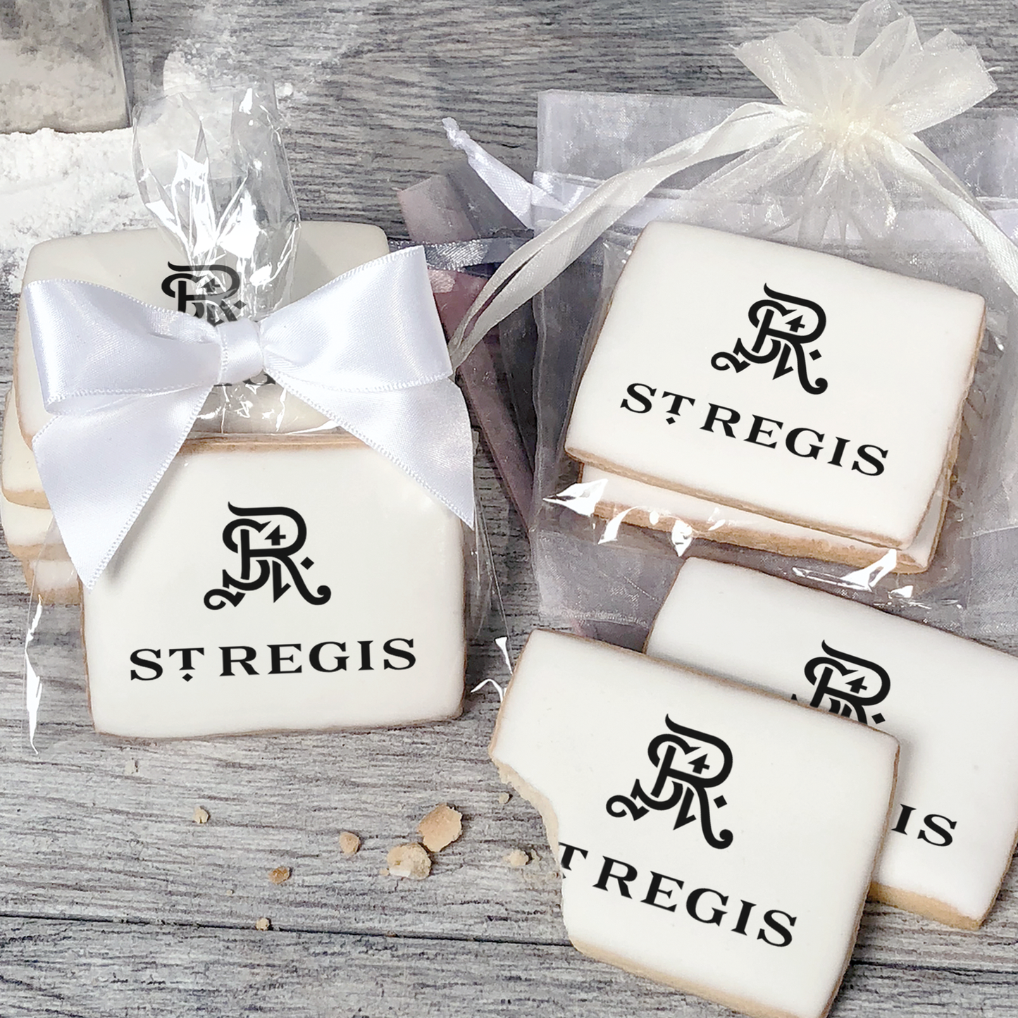 A stack of rectangular butter shortbread cookies with St Regis logo printed directly on a white, light sugar icing. Some cookies are shown in clear packaging with a twist-tie ribbon bow or inside an organza bag.
