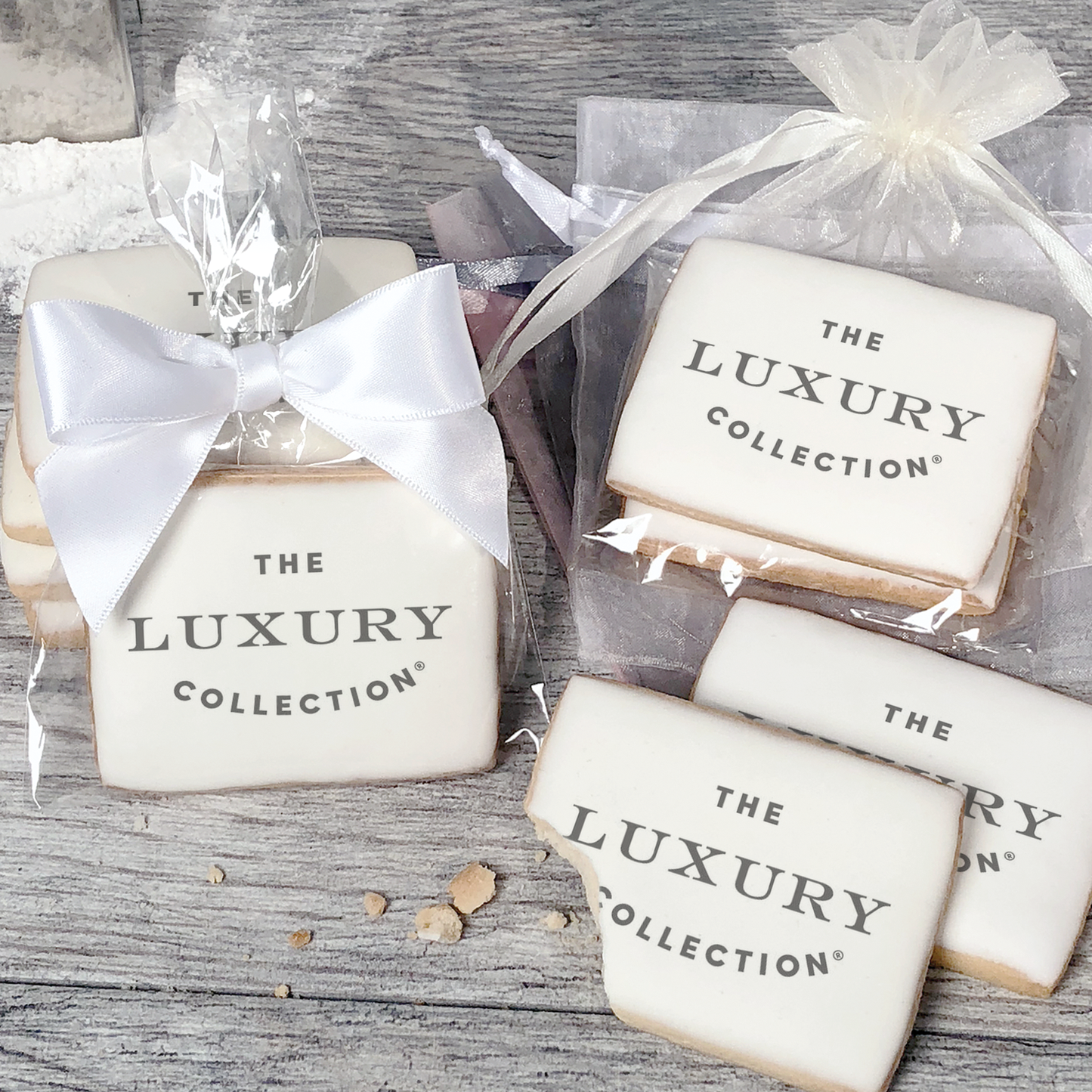 A stack of rectangular butter shortbread cookies with The Luxury Collection logo printed directly on a white, light sugar icing. Some cookies are shown in clear packaging with a twist-tie ribbon bow or inside an organza bag.