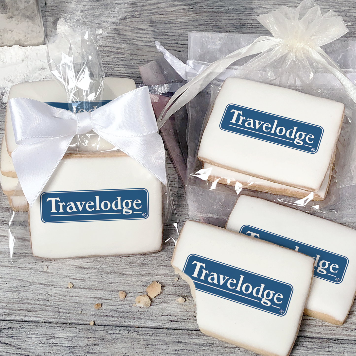 A stack of rectangular butter shortbread cookies with Travelodge logo printed directly on a white, light sugar icing. Some cookies are shown in clear packaging with a twist-tie ribbon bow or inside an organza bag.