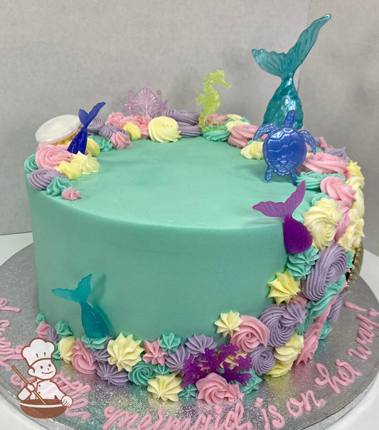 Cake with aqua-colored icing and decorated with plastic mermaid tails and buttercream rosettes in light-pink, yellow, lavender and aqua colors.