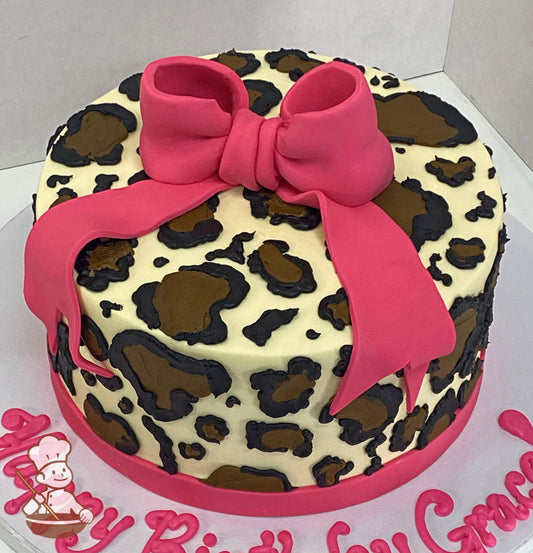 Single tier cake with smooth ivory-colored icing and decorated with hand-piped buttercream cheetah print and a hot pink fondant bow on top.
