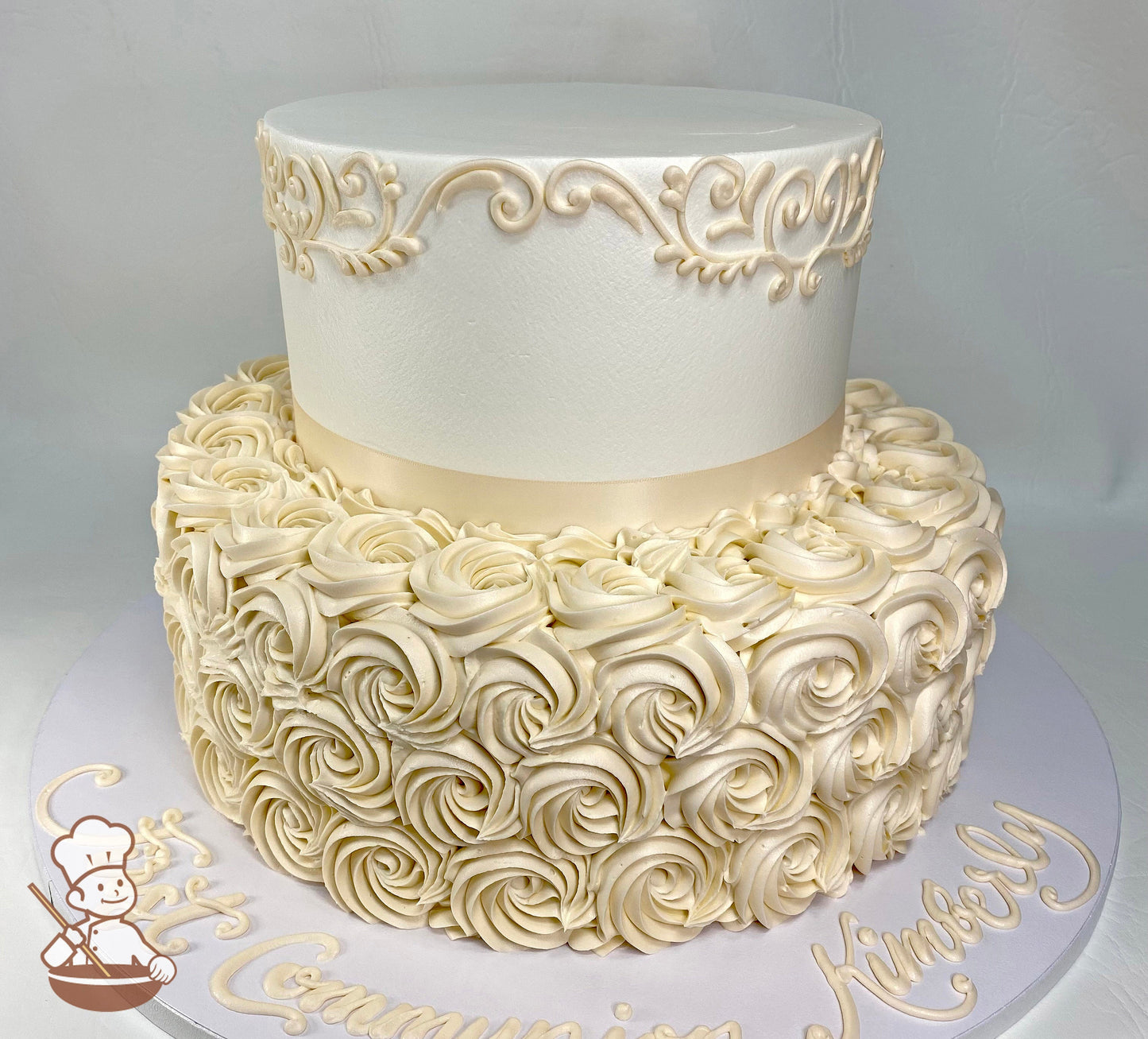 2-tier round cake with buttercream rosettes, has scrolls on cake wall at the top of the tier with an ivory ribbon on the base of the top tier.