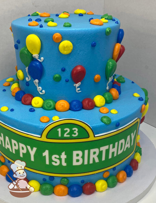 2-tier round cake with sky-blue icing and buttercream balloons and dots in primary colors. Custom happy birthday message printed on bottom tier.