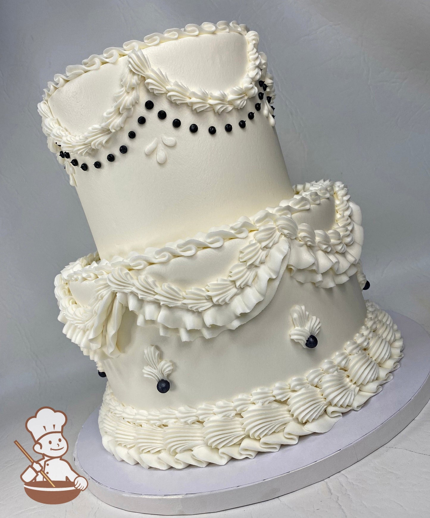 2-tier round cake with smooth white icing and intricate buttercream decorations including large shell-trims and black pearls on the cake walls.