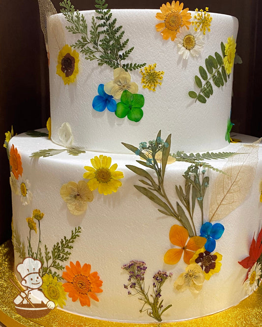2-tier cake with smooth white icing and decorated with dried pressed flowers all over the cake in yellow tones.