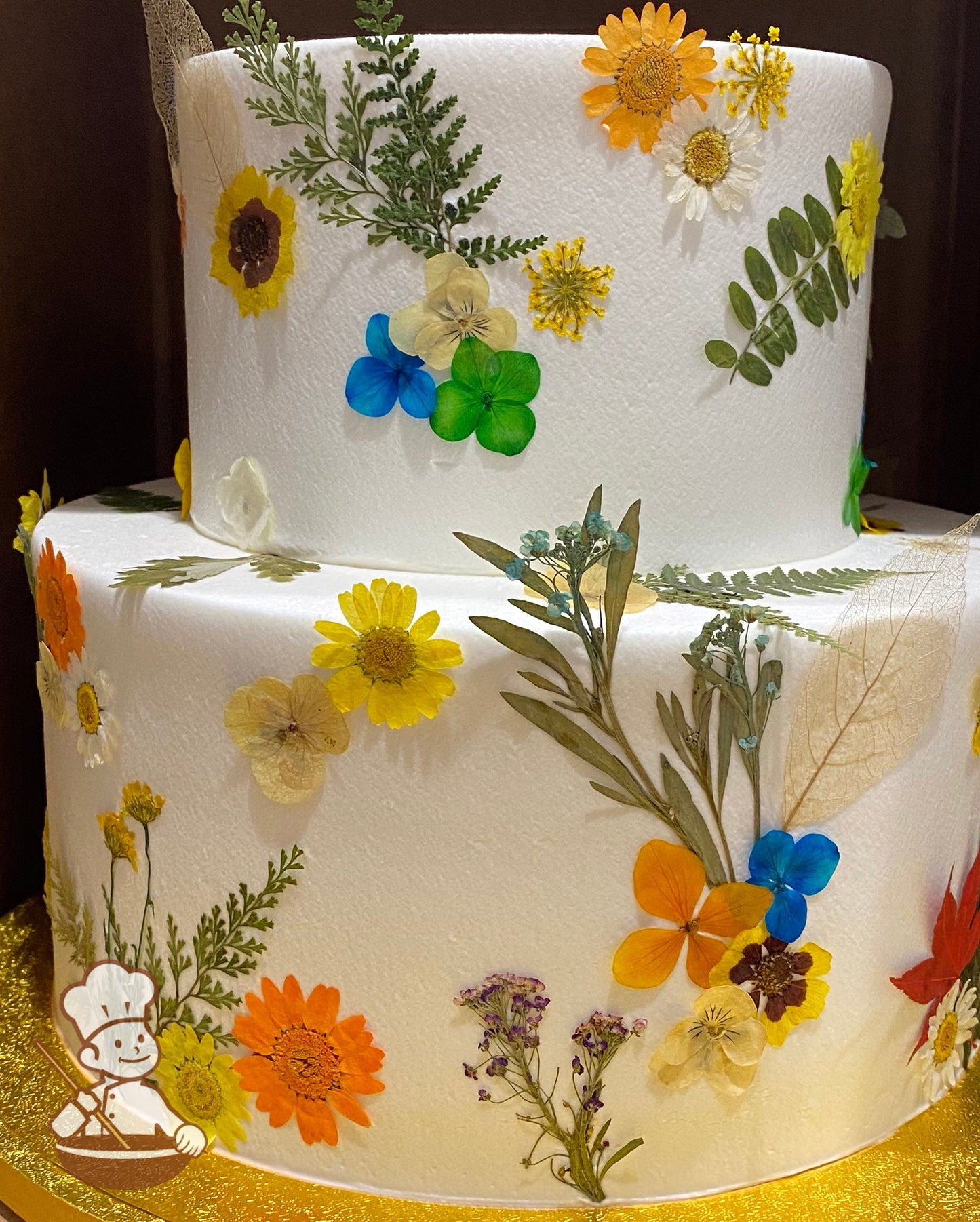 2-tier cake with smooth white icing and decorated with dried pressed flowers all over the cake in yellow tones.