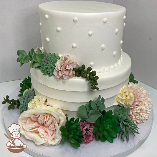 2-tier cake with white icing and decorated with a horizontal texture on the bottom tier and buttercream dots on the top tier and silk flowers.