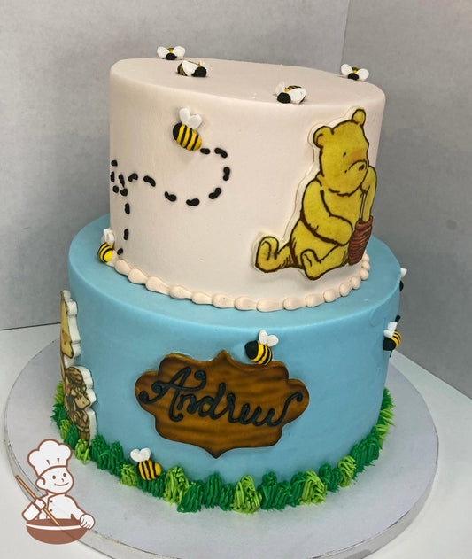 2-tier cake decorated with green buttercream grass, two images of Winnie the Pooh, fondant bees and a custom plaque on the bottom tier.