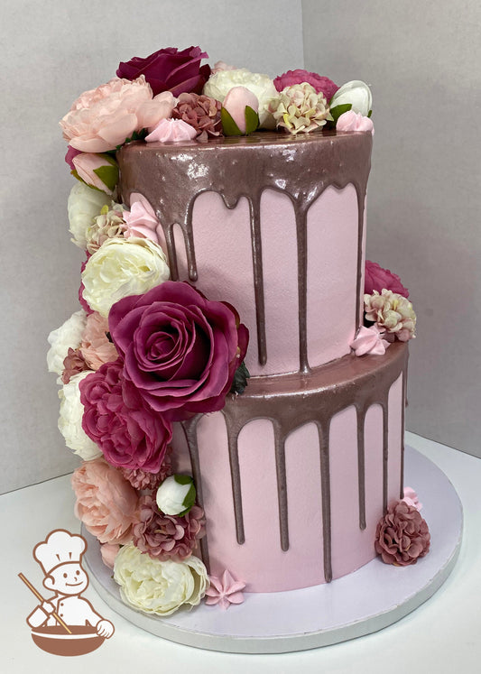 2-tier cake with smooth light-pink icing and decorated with a rose-gold drip and a variety of silk flowers in pink and white colors.