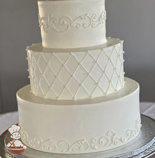 3-tier round cake with white icing, has buttercream scrolls on the bottom and top tier and also has a piped quilt decoration in the middle tier.