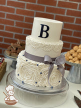 3-tier cake with a white ruffle bottom tier, white scrolls in the middle tier with a gray ribbon bow and a custom fondant letter on the top tier.