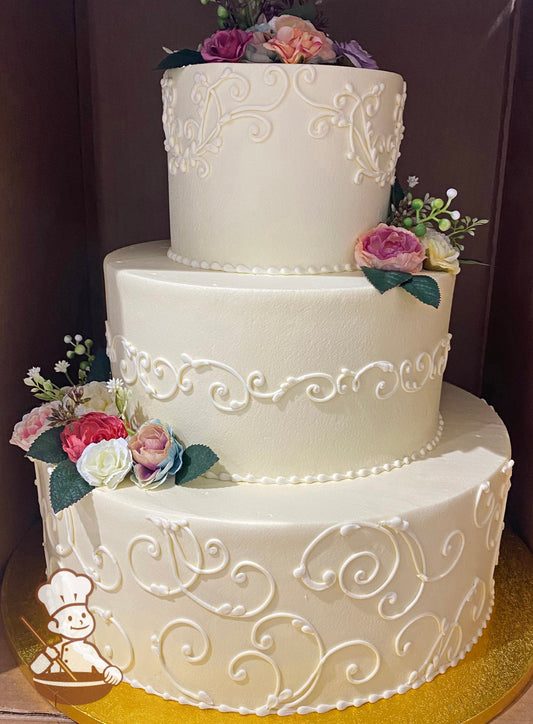 3-tier cake with ivory-tinted smooth icing and decorated with buttercream scrolls and silk flowers in a variety of colors.