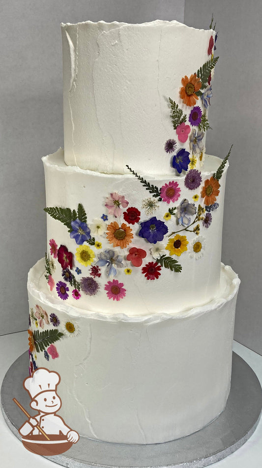 3-tier cake with white icing, decorated with a light texture and dried pressed flowers placed from the top right of the cake to the bottom left.
