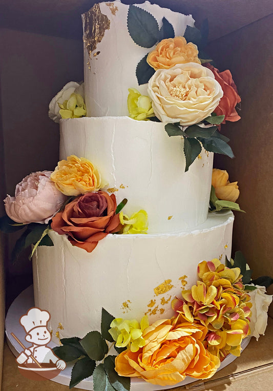 3-tier cake with white icing and decorated with a light texture, gold flakes and silk flowers in orange, yellows, pinks, and cream colors.