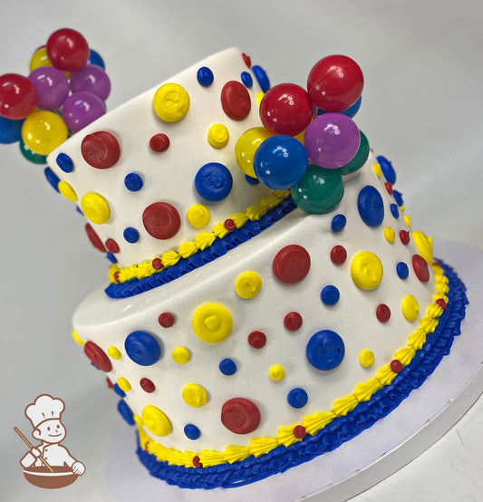 2-tier round cake with buttercream dots on cake walls in primary colors has a plastic bunch of balloons toy added on top tier and bottom tier.
