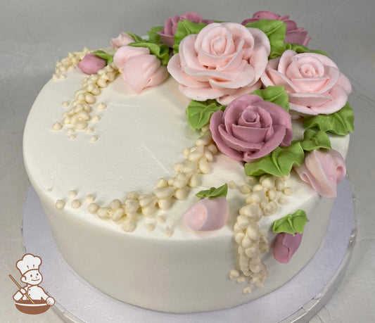 Round cake with white icing and buttercream roses in different shades of pink with green leaves and ivory-colored strings of pearls.