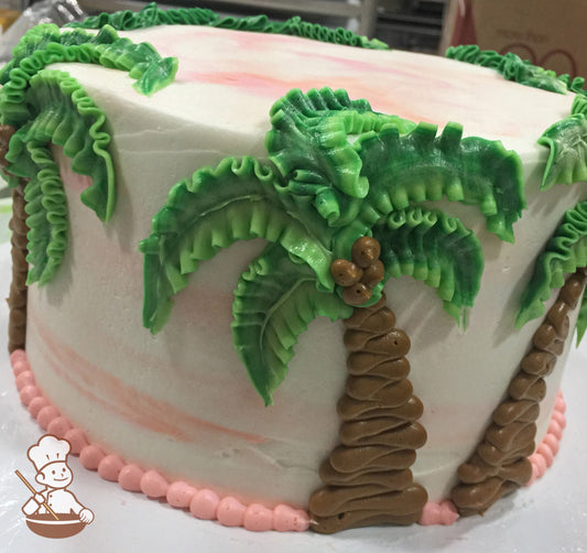Round cake with white icing and smears of peach-colored watercolor decorated with buttercream palm trees and a peach-colored beaded trim.