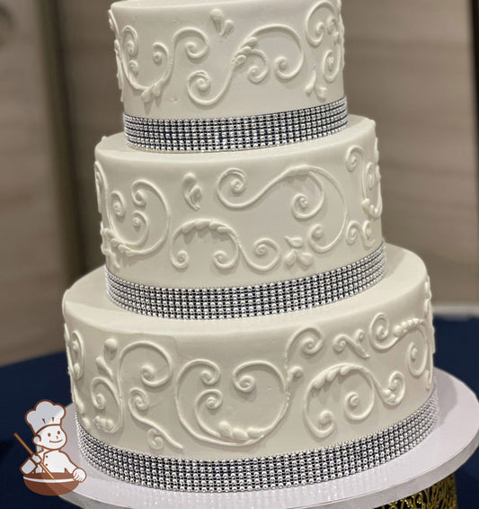 3-tier round cake with white icing and a crystal bling band on bottom of each tier with white buttercream scroll decorations on the cake walls.