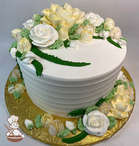 Single tier round cake with white icing and a horizontal texture. Cake has a variety of buttercream florals in pastel-yellow and white colors.