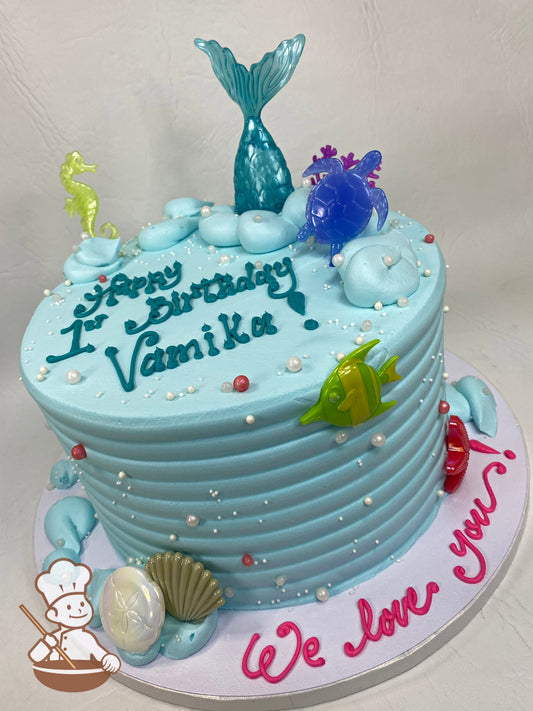 Single tier round cake with light blue icing and a horizontal texture, it also has plastic mermaid toys in jewel tones and white pearls all over.