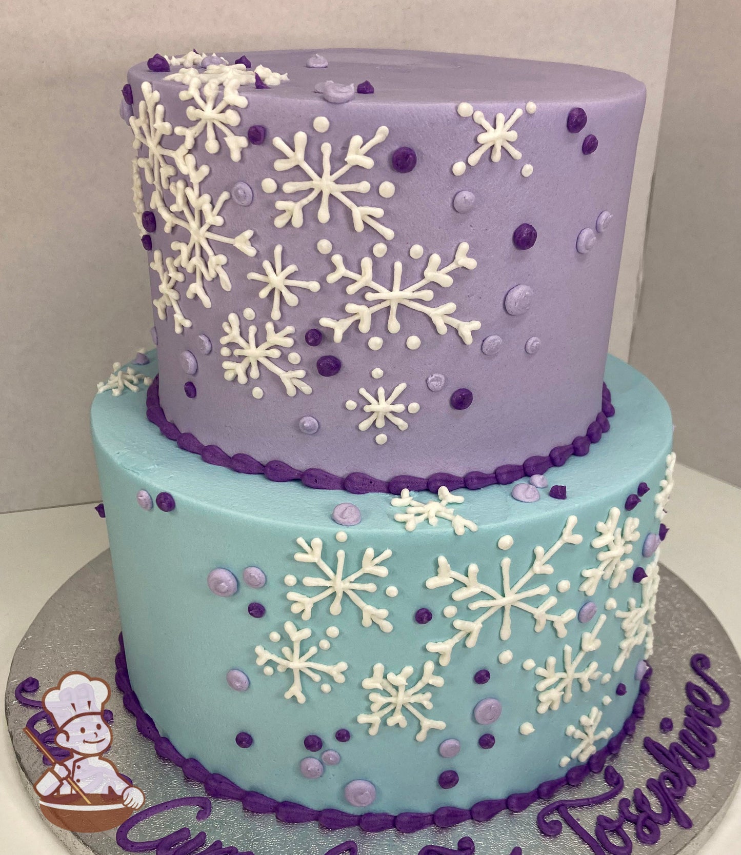 2-tier round cake with light blue icing on bottom tier and lavender icing on top tier with buttercream white snowflakes on cake walls with dots.