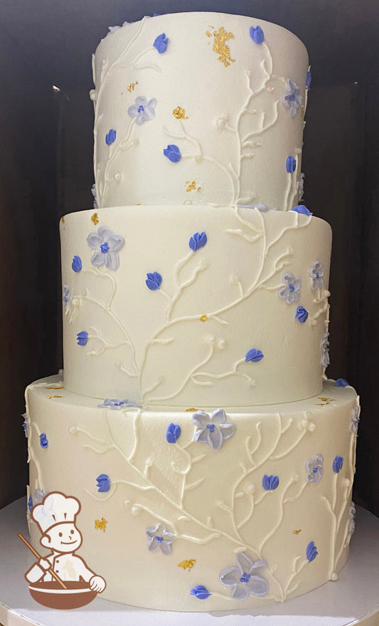 3-tier round cake with white smooth icing and hand-piped periwinkle flowers and long white buttercream vines with a touch of gold foil flakes.
