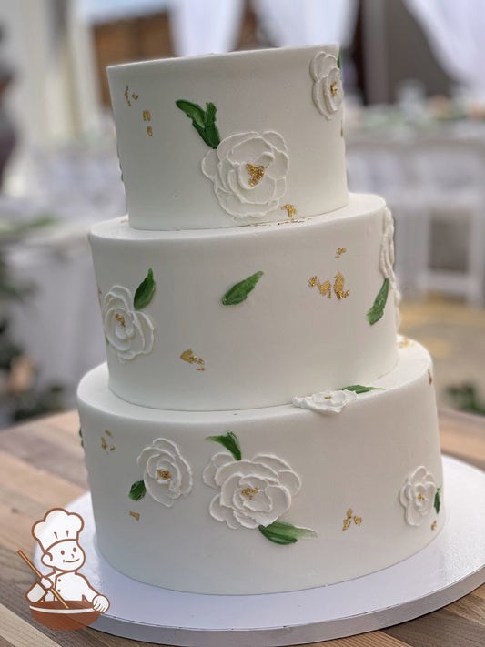 3-tier cake with white icing and hand-crafted flowers in white and green accents with gold foil flakes placed elegantly on the cake tiers.