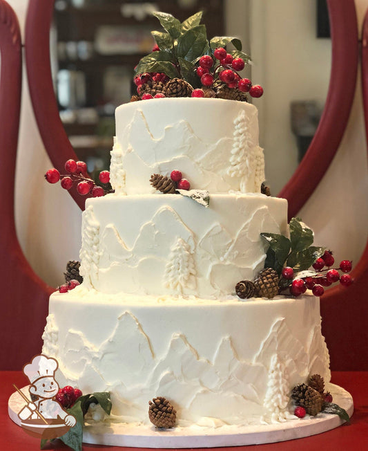 3-tier cake with white icing, decorated with buttercream mountains and trees on the cake walls and fake brown pinecones and red cranberries.