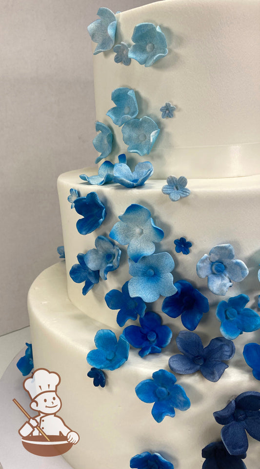 3-tier cake with smooth white icing, decorated with sugar blossoms in different shades of blue cascading on the cake.