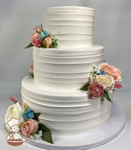 3-tier cake with white icing and decorated with a horizontal texture and silk flowers in a variety of colors.