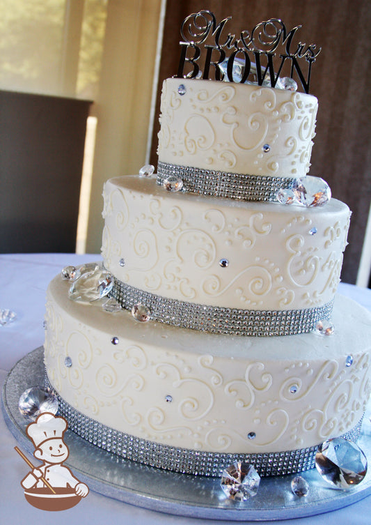 3-tier cake with smooth white icing decorated with white buttercream scrolls, a rhinestone band on the bases and diamonds on the tiers.