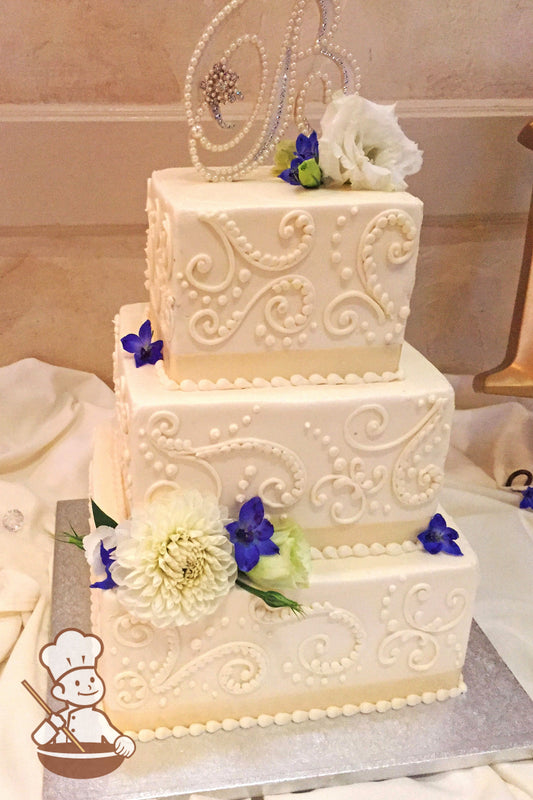 3-tier cake with smooth white icing and white buttercream scrolls and a cream-colored ribbon on the base of each tier with fresh flowers.