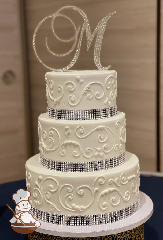 3-tier cake with smooth white icing decorated with white buttercream scrolls, a black satin ribbon with a rhinestone band on top of it.