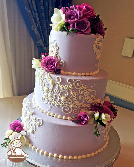 3-tier cake with lavender-tinted smooth buttercream with white buttercream scrolls and purple fresh flowers.