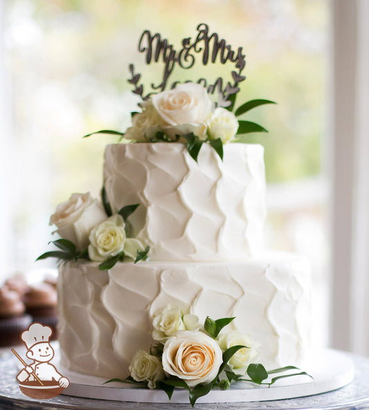 2-tier cake with white icing and decorated with a vertical wavy texture and white and cream colored fresh flowers.